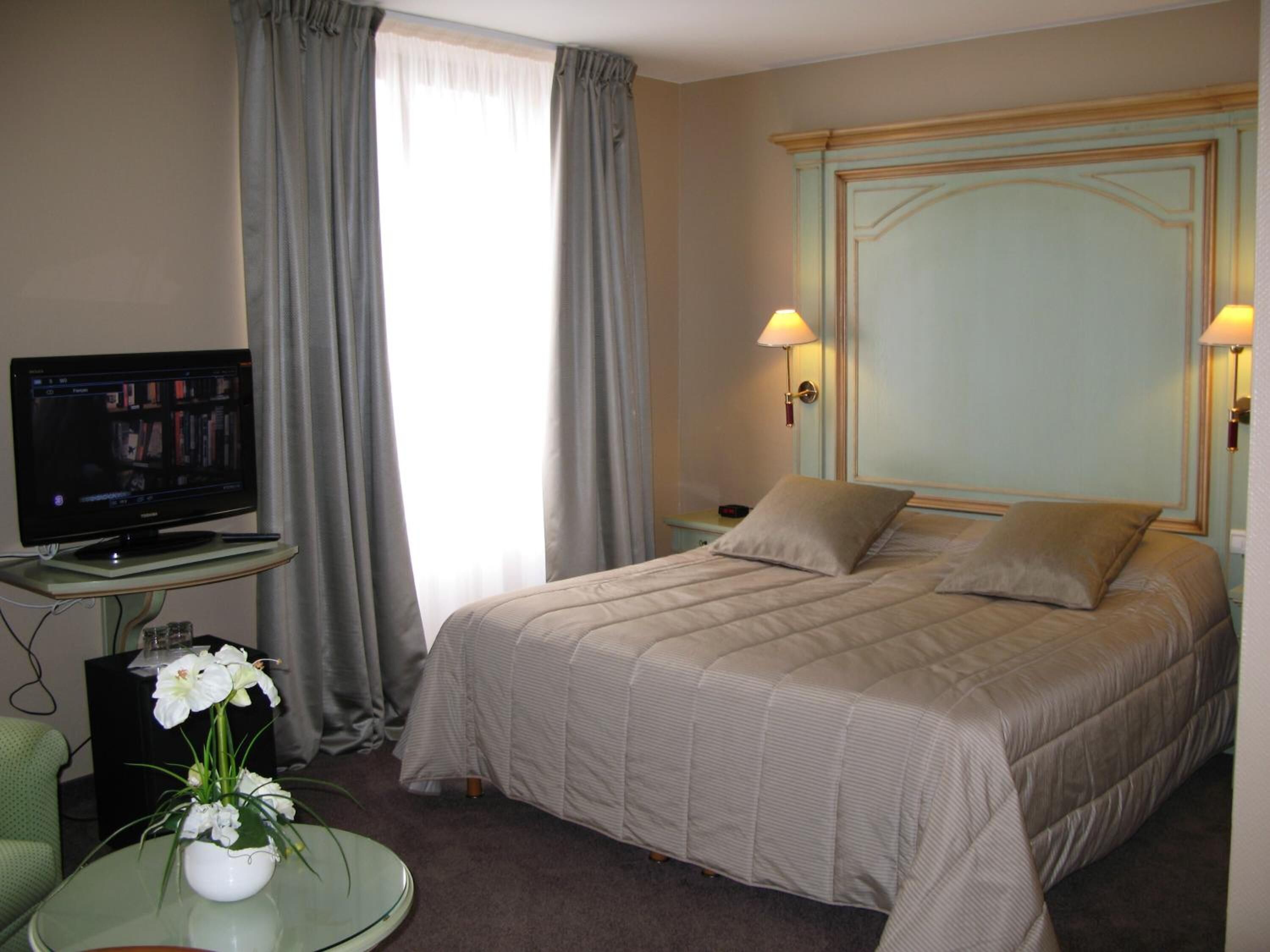 Habitación de hotel con una cama, un televisor sobre un soporte, una mesita de noche, una lámpara y un jarrón con flores.