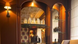 Uma área de estar acolhedora com sofá, banqueta e poltrona, uma mesa pequena com um copo, e uma parede com prateleiras exibindo garrafas.