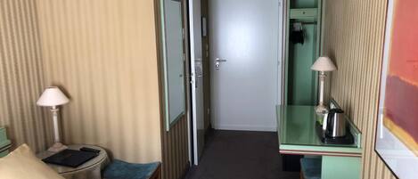 Habitación de hotel con cama, escritorio con lámpara, taburete pequeño, cubo de basura y puerta que da a otra habitación.