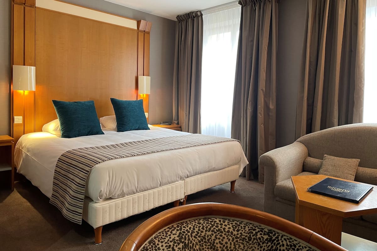 Habitación de hotel con una cama grande, dos lámparas de noche, cabecera de madera y vista a la ciudad a través de cortinas.