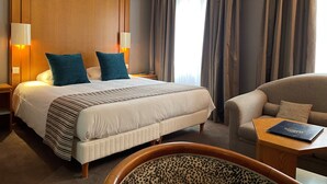 Quarto de hotel com uma cama grande, duas luminárias de cabeceira, um painel de cabeceira de madeira e vista para a cidade através das cortinas.