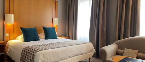 Habitación de hotel con una cama grande, dos lámparas de noche, cabecera de madera y vista a la ciudad a través de cortinas.