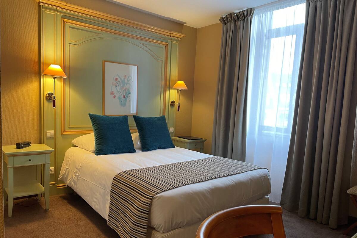 Una habitación de hotel con cama, dos mesitas de noche, una silla y un ventanal con cortinas.