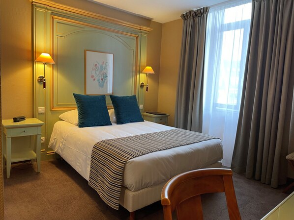 Una habitación de hotel con cama, dos mesitas de noche, una silla y un ventanal con cortinas.