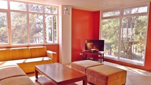 A living room with a sofa, ottoman, and coffee table, a television, and large windows with a view of trees. - Arc Residences (Baguio)