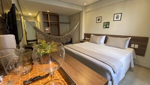 Una habitación de hotel con una cama grande, una mesita de noche de madera, una mesa de vidrio con copas de vino y una hamaca al fondo.