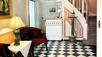 Una habitación con piso a cuadros, una escalera, una mesa con lámpara y un cuadro en la pared.