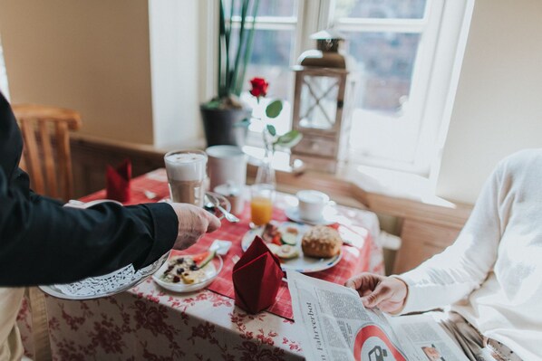 Una escena de desayuno con una mesa preparada para dos personas, que incluye un periódico, una servilleta roja y diversas opciones para el desayuno, como tostadas, fruta y una bebida.