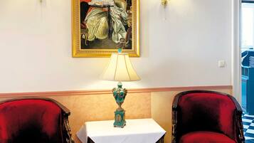 Una habitación con piso a cuadros, dos sillones tapizados en rojo, una mesa con mantel blanco, una lámpara turquesa y un cuadro enmarcado en la pared.