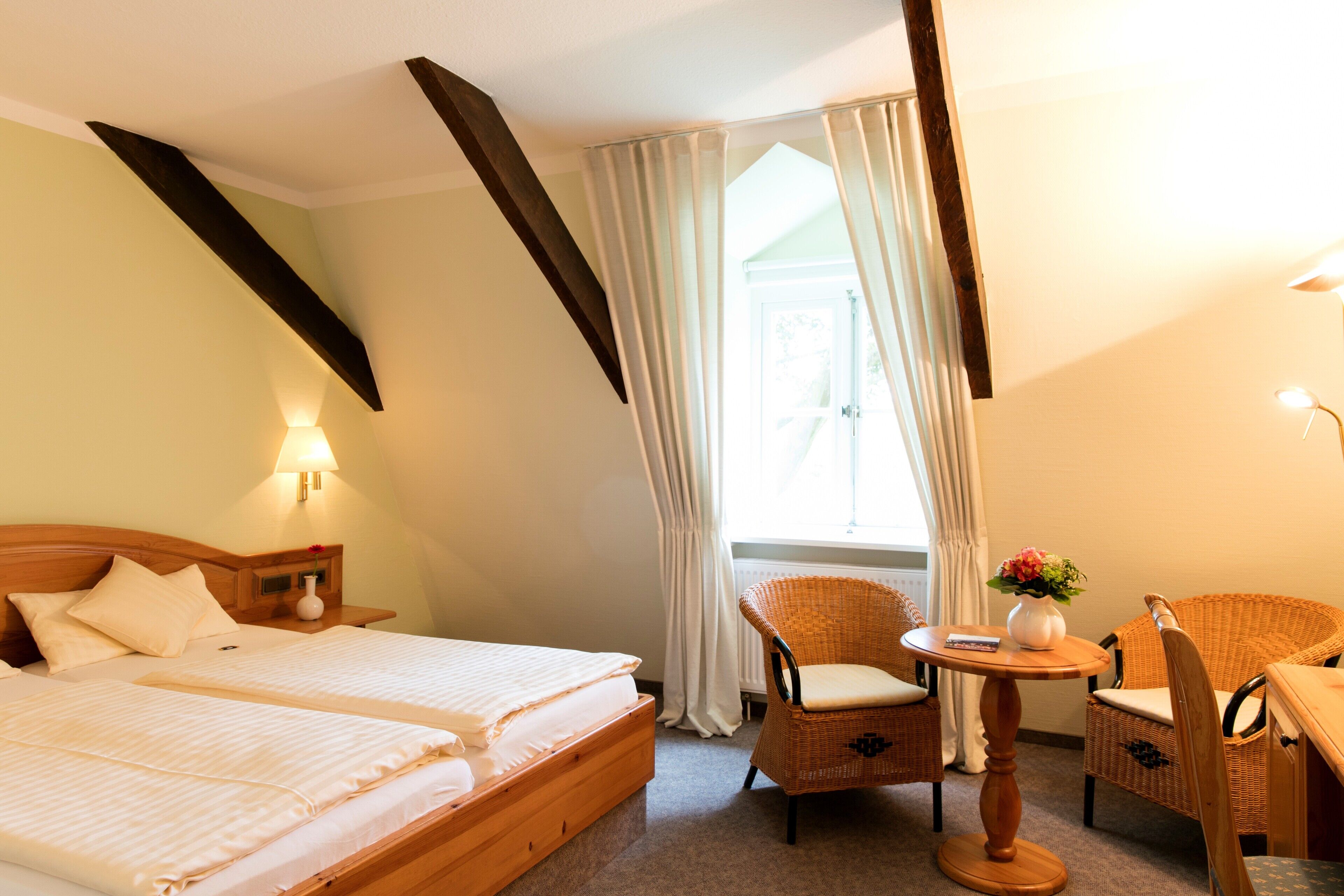 A hotel room with two beds, a sitting area with wicker chairs, a small table with a vase, and a window with curtains.