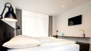 Un dormitorio moderno con una cama grande, una mesita de noche, un televisor en la pared y una ventana con cortinas.