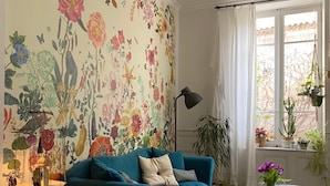 Un salón con papel tapiz floral, un sofá azul verdoso, suelo estampado y una ventana con plantas.