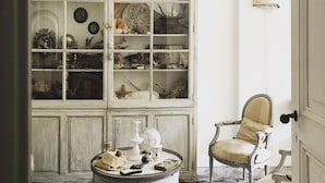 Una habitación con una estantería blanca llena de libros y objetos decorativos, una mesa redonda con un globo terráqueo y una vela, y una silla antigua.