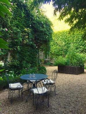 Un patio jardín con una mesa redonda y cuatro sillas, rodeado de follaje denso y una jardinera de madera.