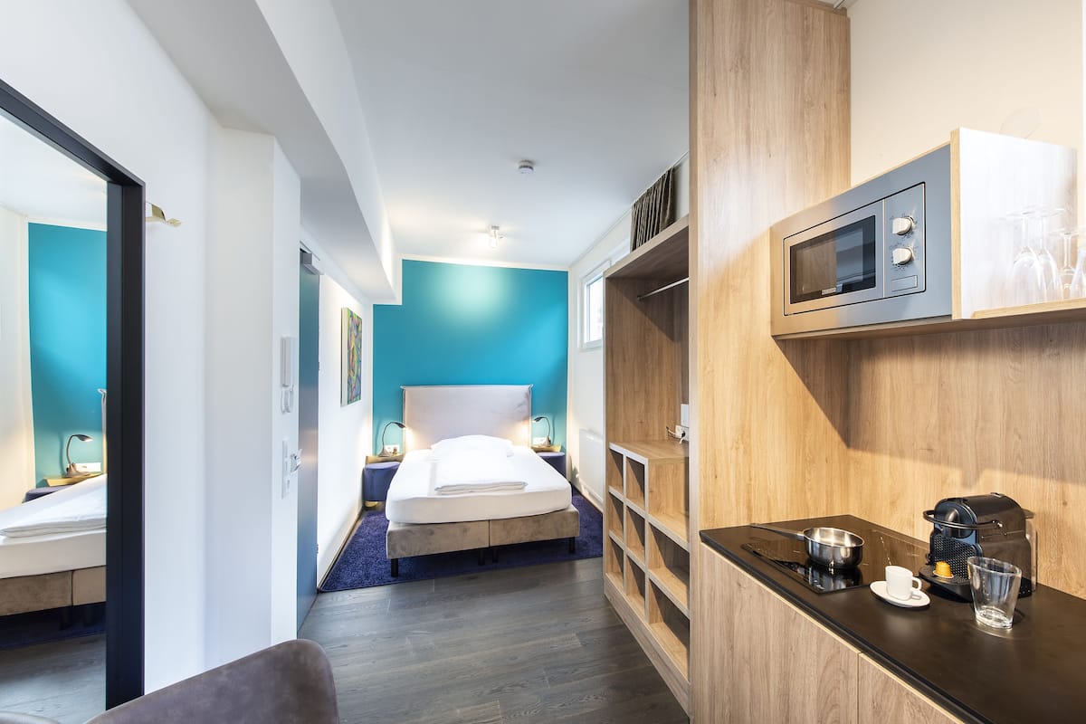 A modern hotel room with a bed, a kitchenette, and a view of the hallway.