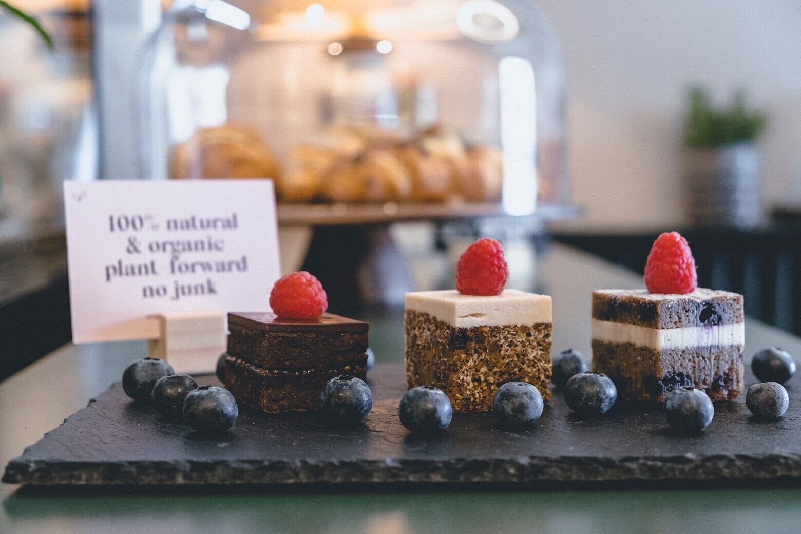 Trois desserts carrés présentés sur une ardoise, chacun surmonté d’une framboise et entouré de myrtilles.