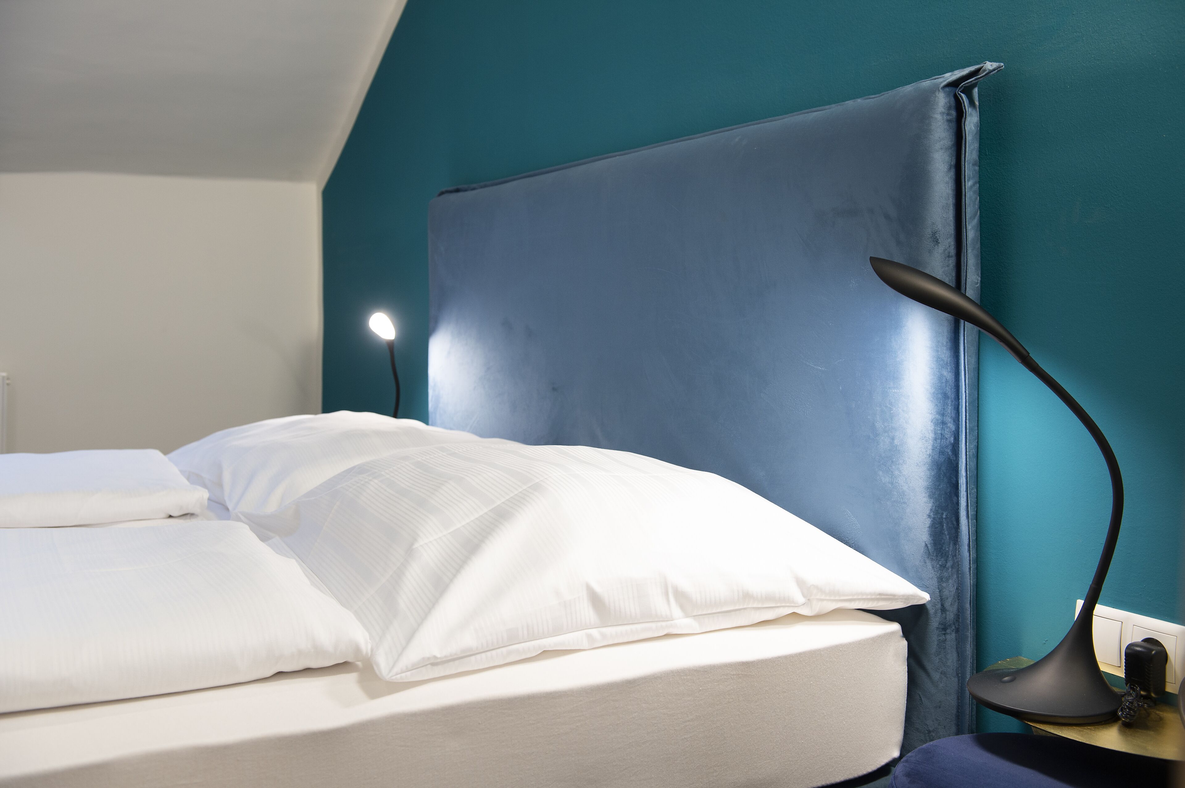 A neatly made bed with white linens in a room with teal walls and a modern lamp on a bedside table.