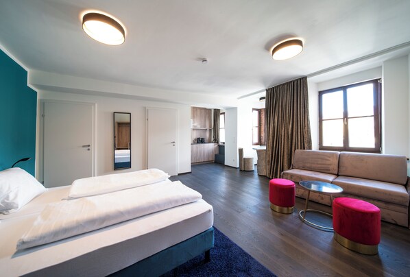 A modern hotel room with a bed, a sofa, two red ottomans, a small round table, and a view of the kitchen and living area.