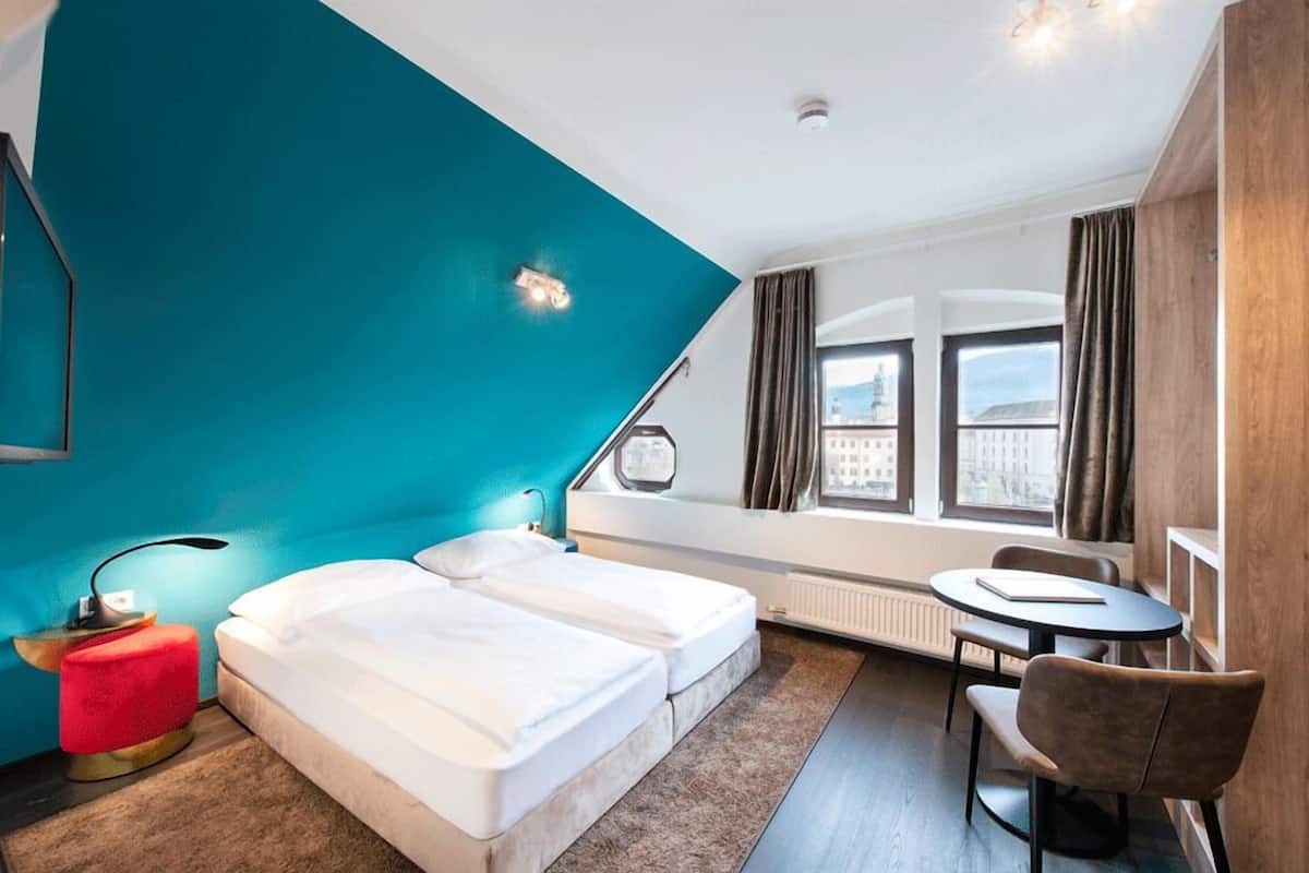 A hotel room with a teal accent wall, two beds, a small round table with a chair, and a window with curtains.