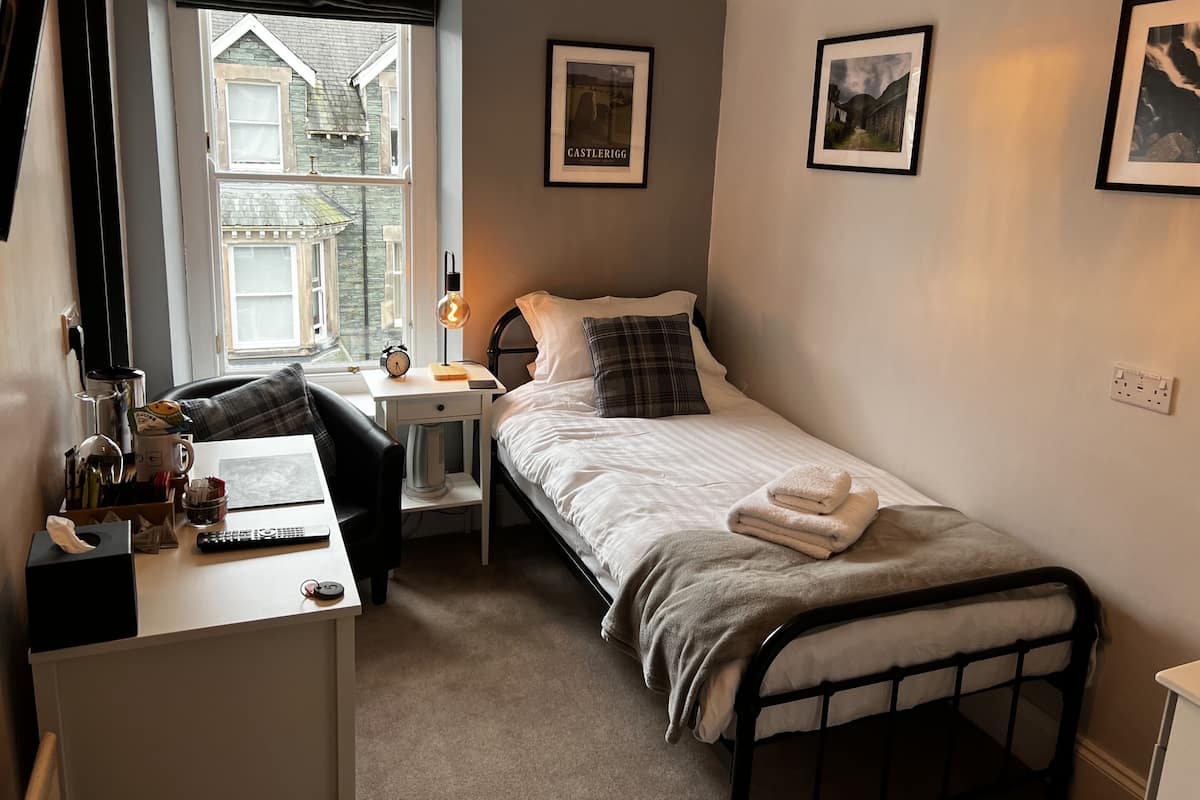 Een slaapkamer met een bed, nachtkastje, lamp en een raam met uitzicht op een gebouw.