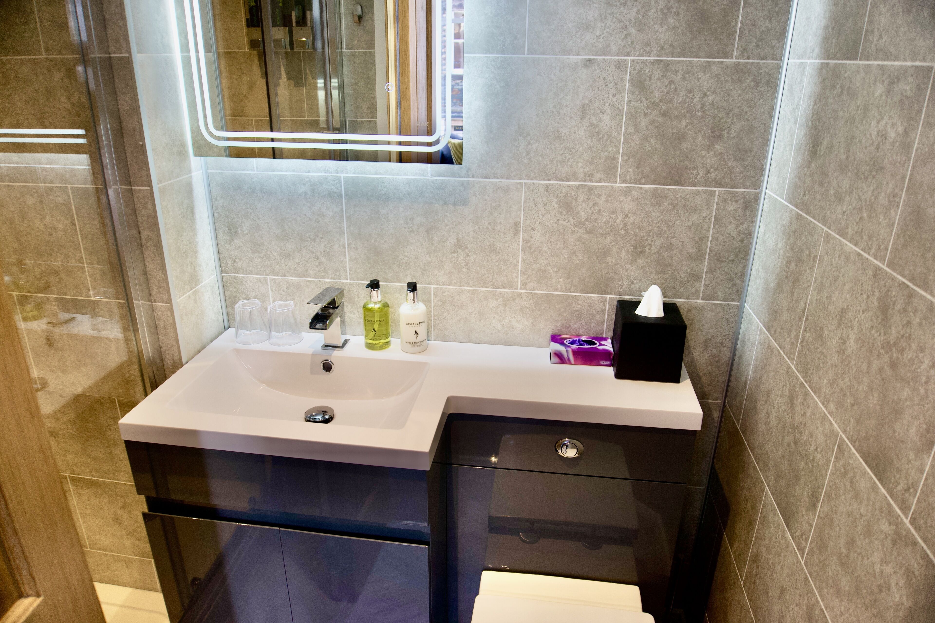 Un baño moderno con un lavabo blanco, dos botellas de jabón líquido, una caja de pañuelos negros y un espejo con una tira de luz.