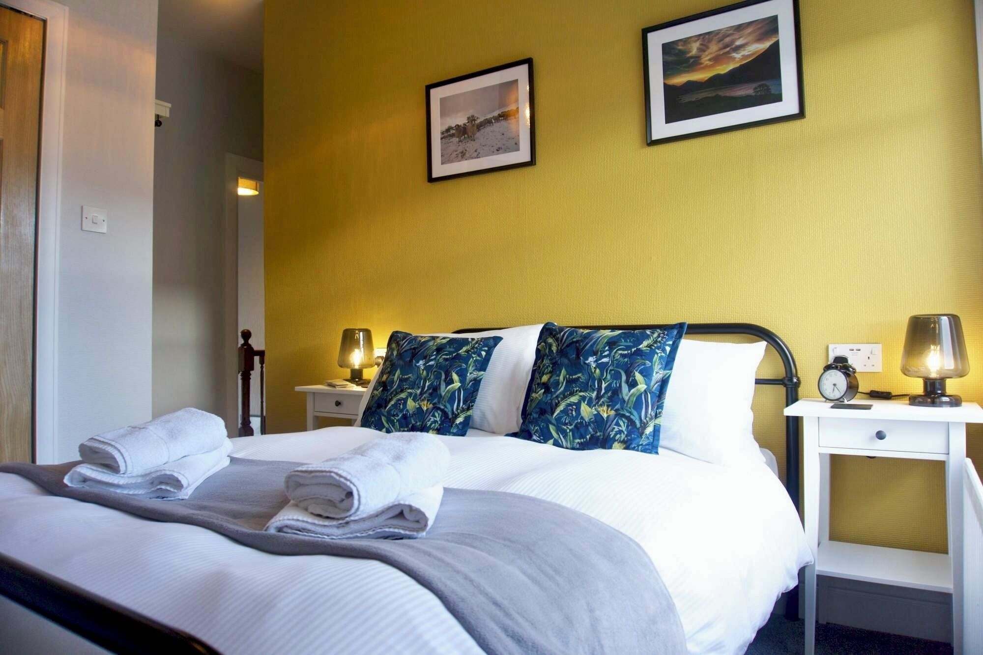 Habitación de hotel con una cama, dos almohadas, una mesita de noche con lámpara y un mueble auxiliar con reloj.