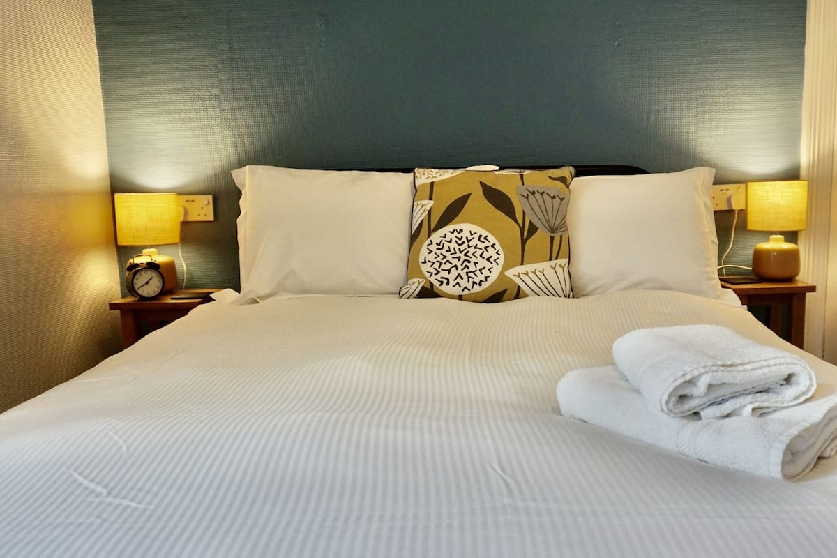 Een net opgemaakt bed met witte lakens, twee bedlampen en een decoratief kussen met een bladmotief.