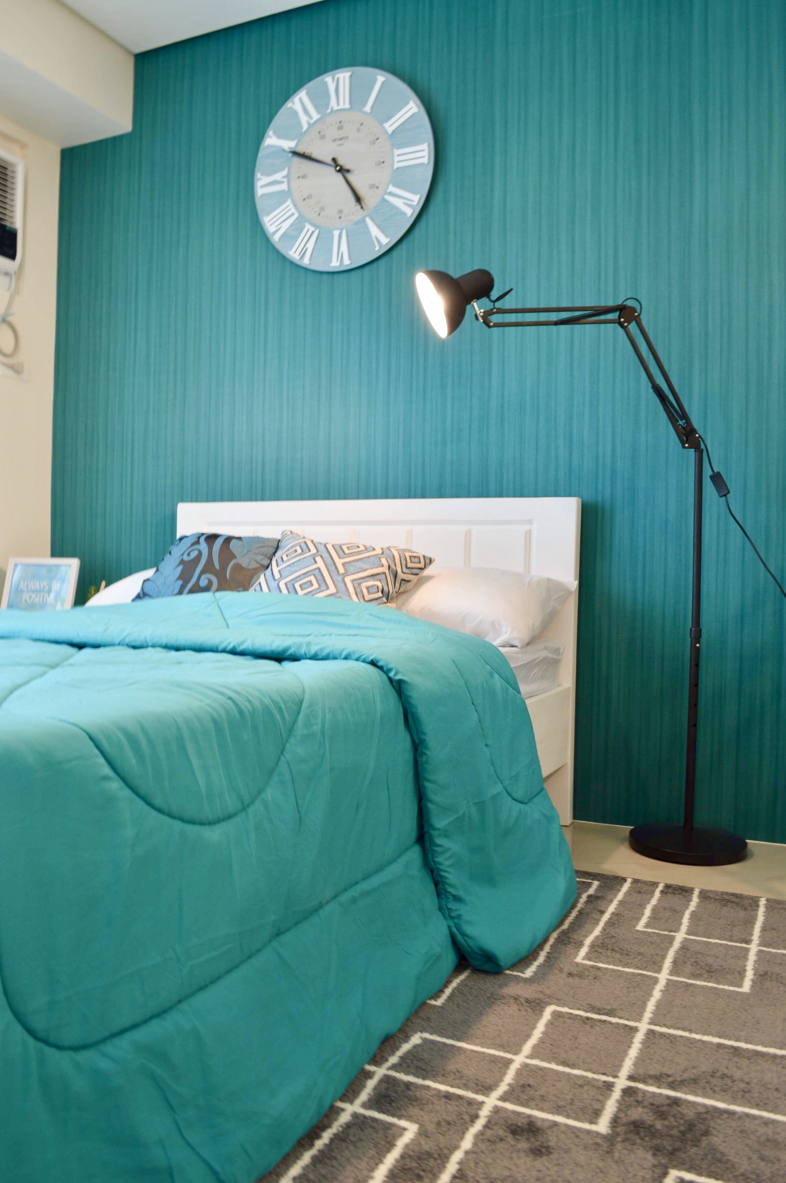 A bedroom with teal walls, a white headboard, a teal bedspread, a round wall clock, and a floor lamp.