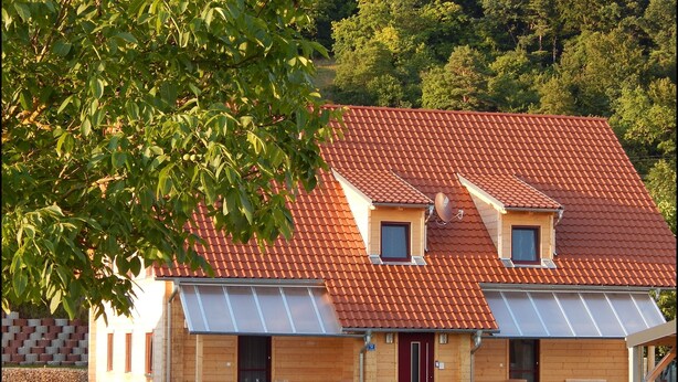 Une maison en bois à deux étages, avec un toit recouvert de tuiles rouges, une véranda et un petit jardin agrémenté de meubles d’extérieur.