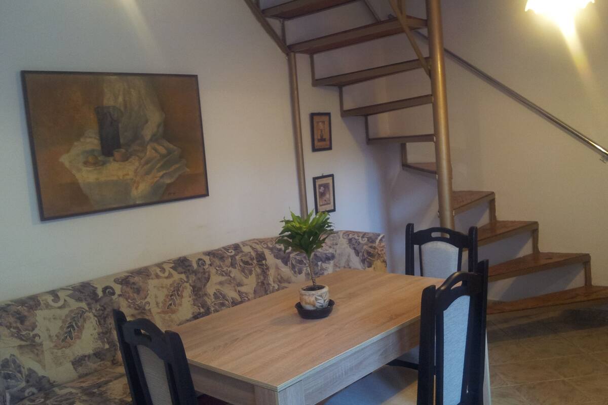 A dining area with a wooden table, two chairs, a bench, and a potted plant. A staircase with a metal railing is visible in the background.