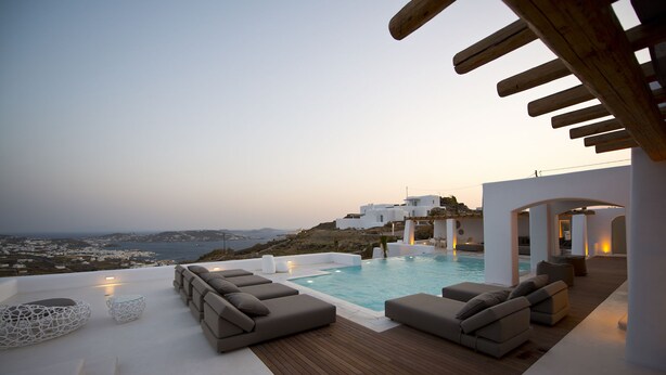 Un área de piscina exterior moderna con vista al mar y edificios blancos al fondo.
