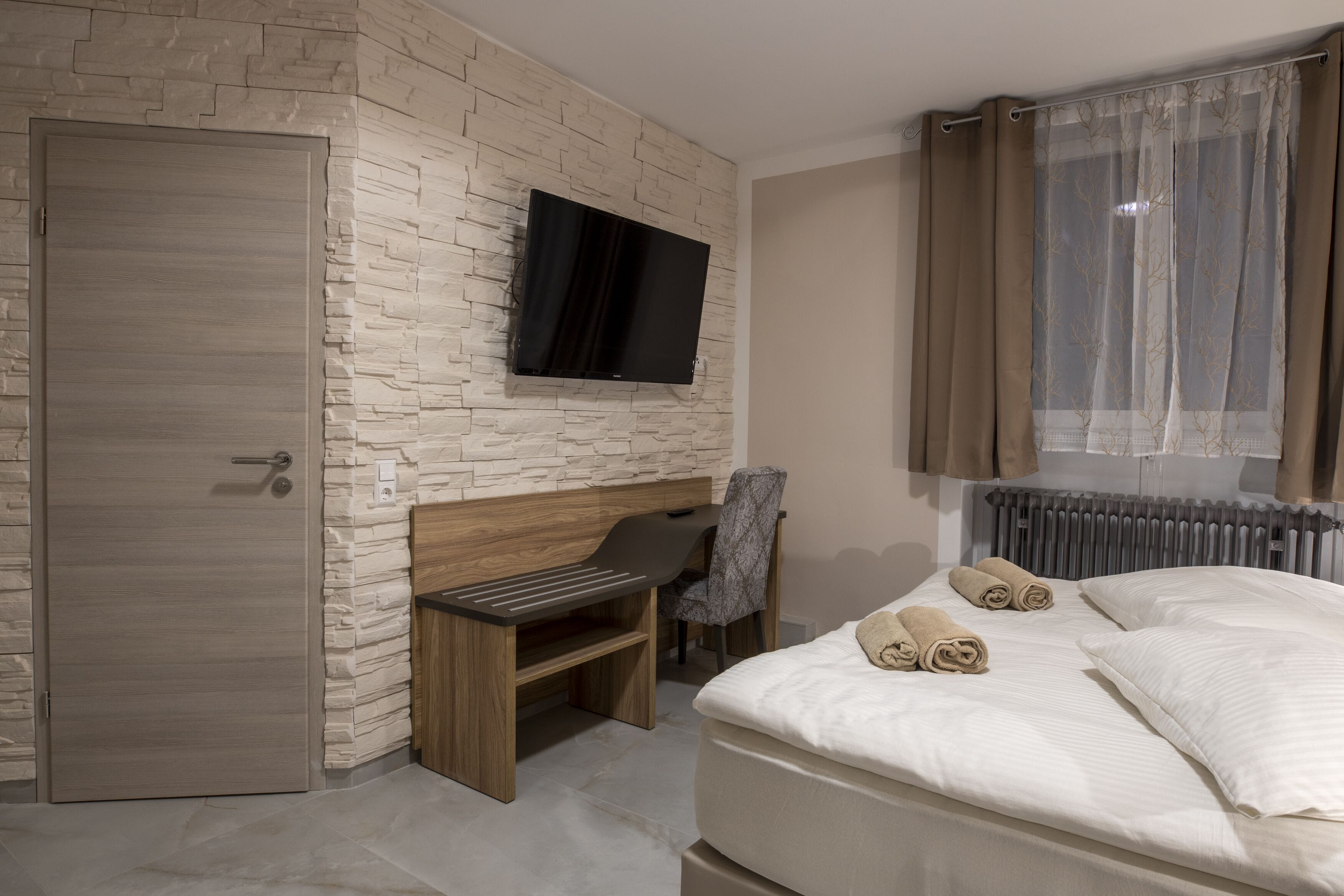 Une chambre d’hôtel avec un mur en pierre, un téléviseur à écran plat, un lit avec des serviettes pliées, un bureau avec une chaise et une fenêtre avec des rideaux.