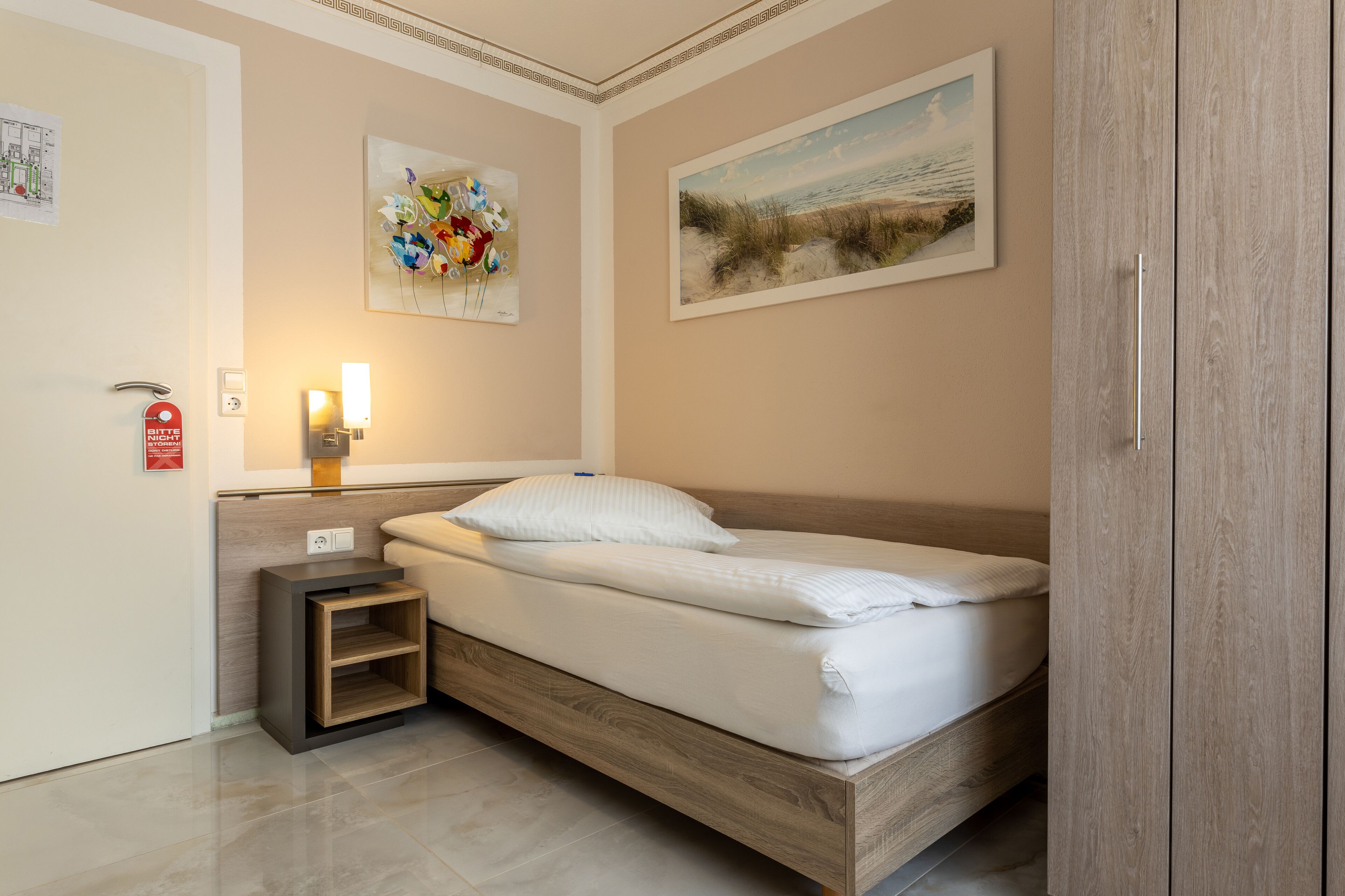 Een netjes opgemaakt bed met hoofdbord, een nachtkastje met lamp en twee ingelijste kunstwerken aan de muur.