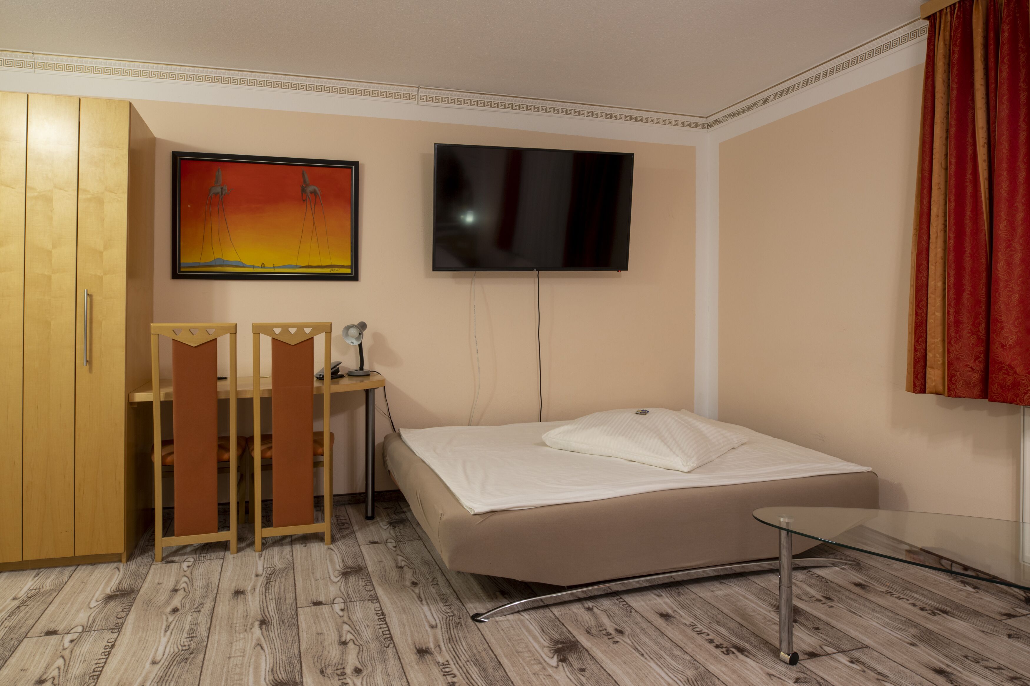 A hotel room with a bed, a desk with a chair, a wardrobe, a TV, and a painting on the wall.