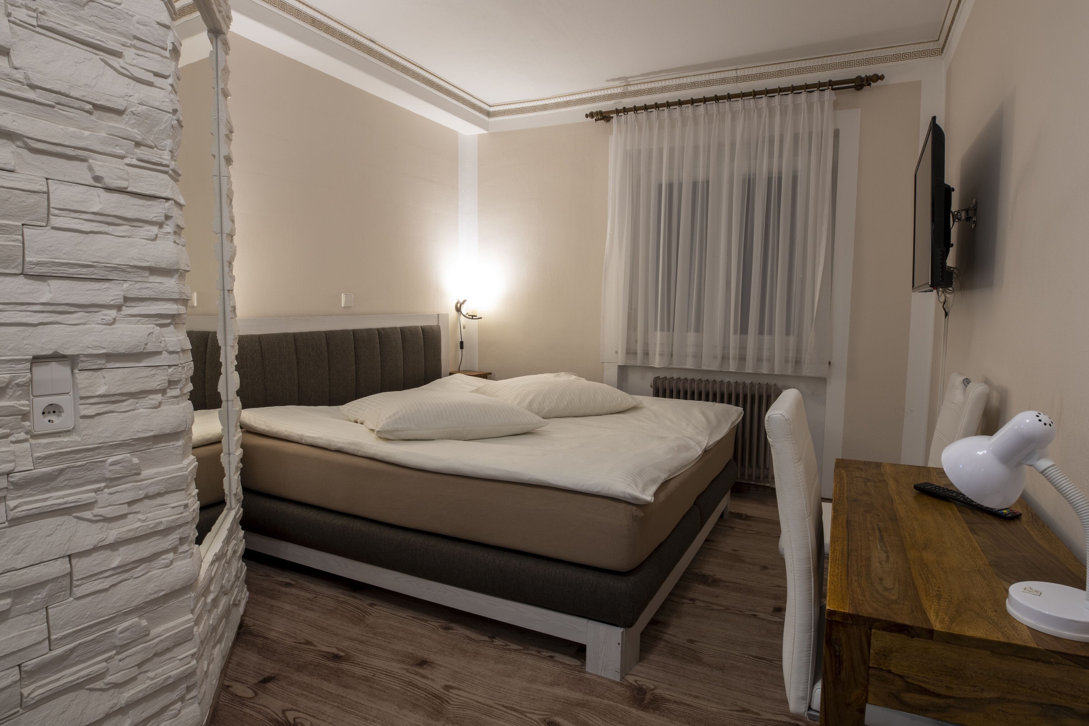 Une chambre à coucher avec un lit, une table de chevet avec une lampe, un téléviseur fixé au mur et une fenêtre avec des rideaux.