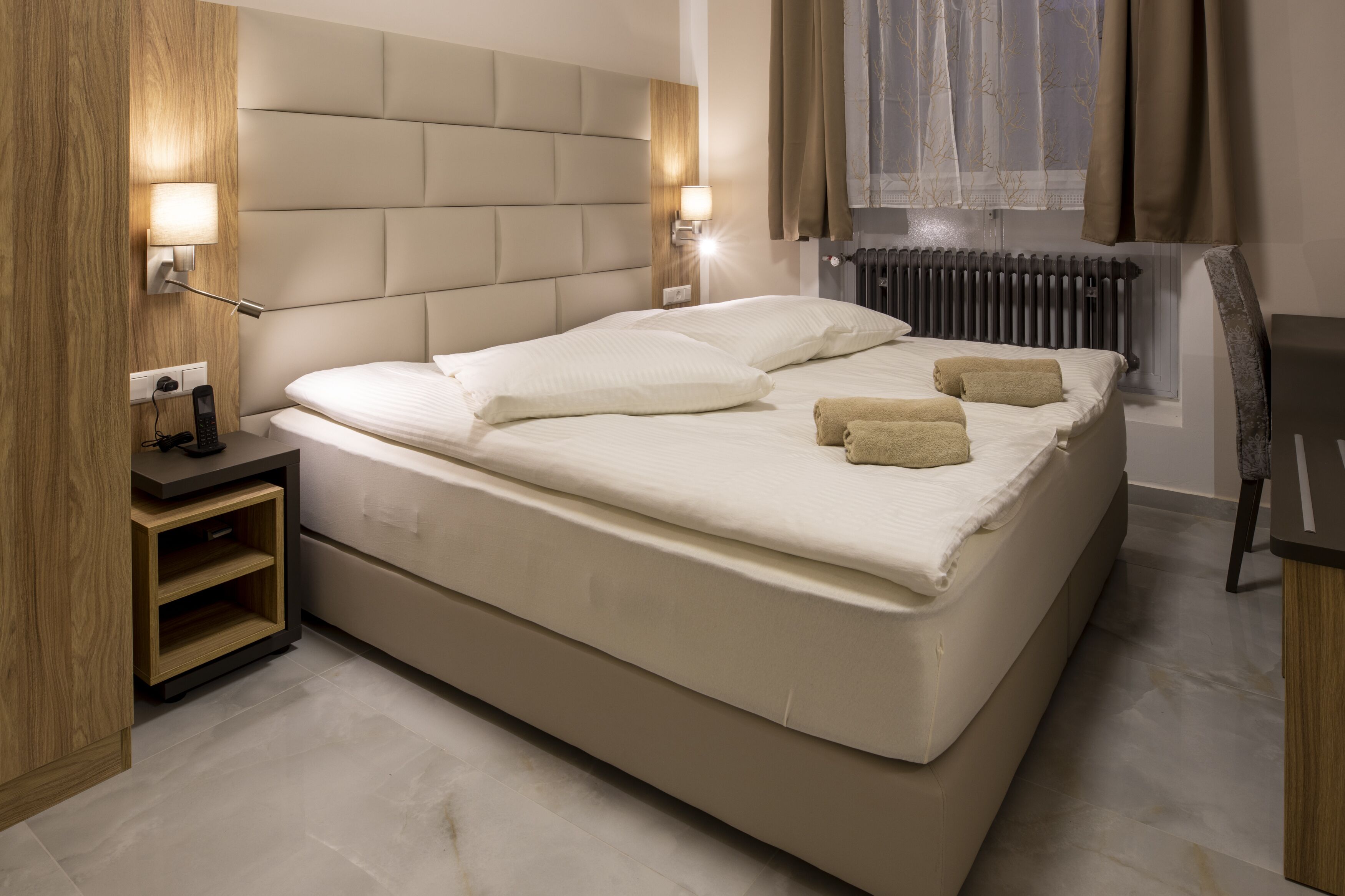 A modern hotel room with a large bed, bedside lamps, a wooden nightstand, and a view of the outdoors.