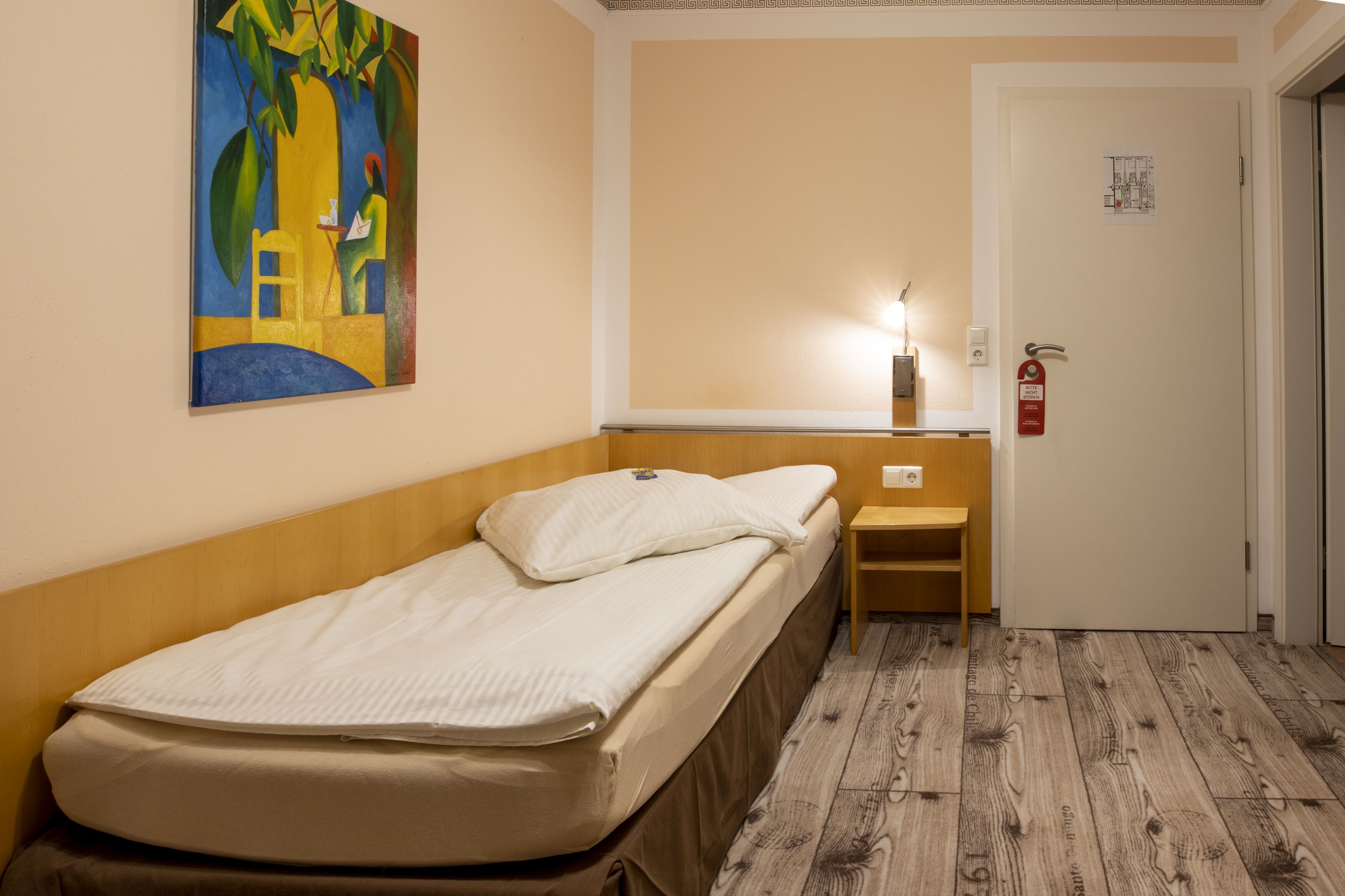 A single bed with a wooden headboard, a bedside table, a nightstand, a wall-mounted lamp, and a painting.