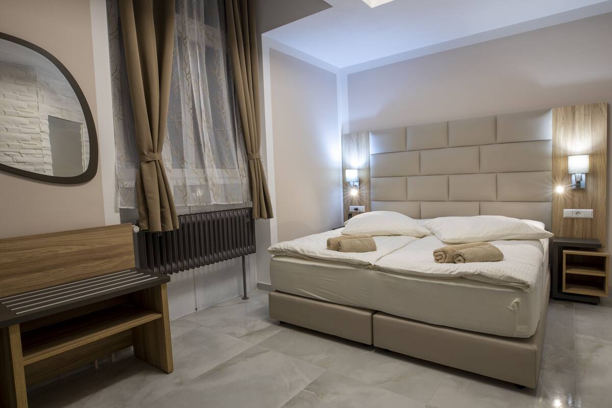 A modern bedroom with a large bed, a wooden bench, a radiator, and a wall-mounted mirror.
