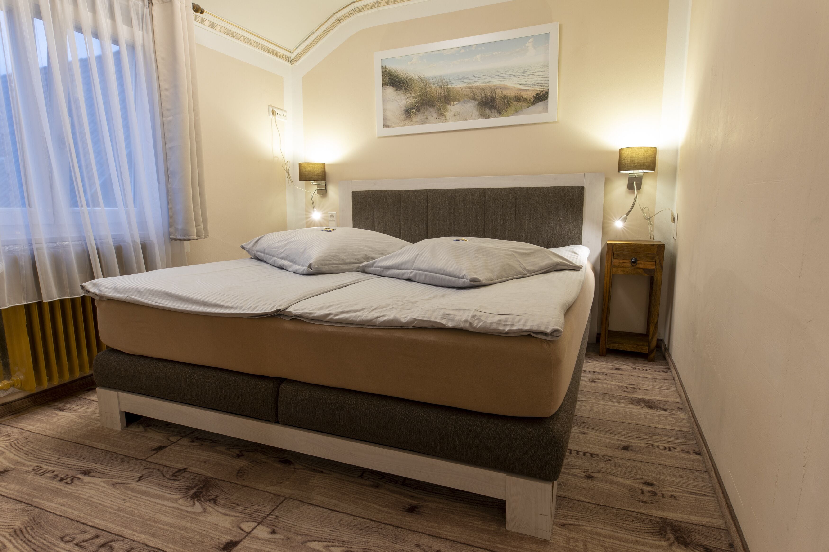 A bedroom with a bed, two bedside tables, a nightstand, a painting on the wall, and a window with curtains.