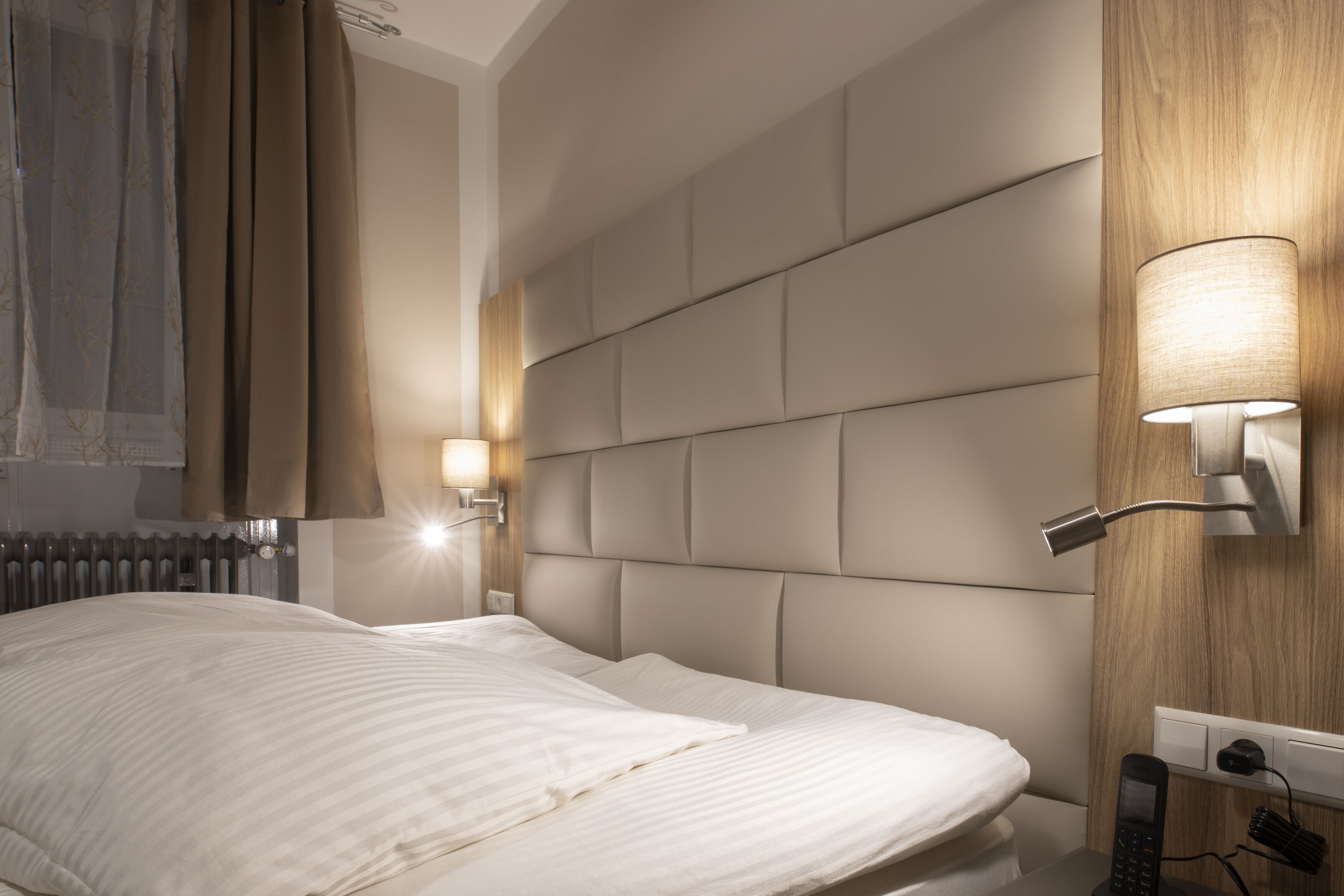 A modern hotel room with a large bed, a headboard with a light panel, a bedside table with a lamp, and a phone.
