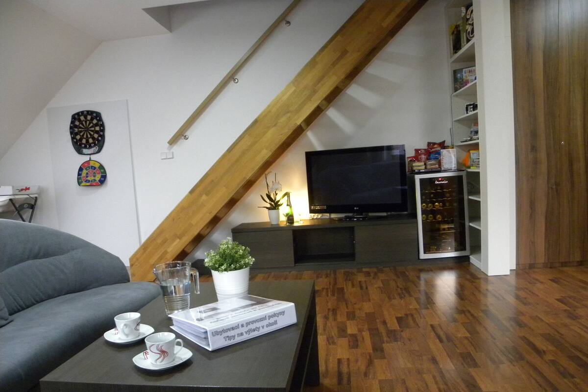A living room with a grey sofa, a coffee table with cups and a book, a TV, and a wooden ceiling beam.