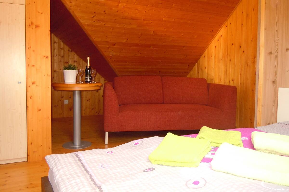 A cozy room with a wooden ceiling, a red sofa, a small table with a bottle and glasses, and a bed with neatly folded towels.