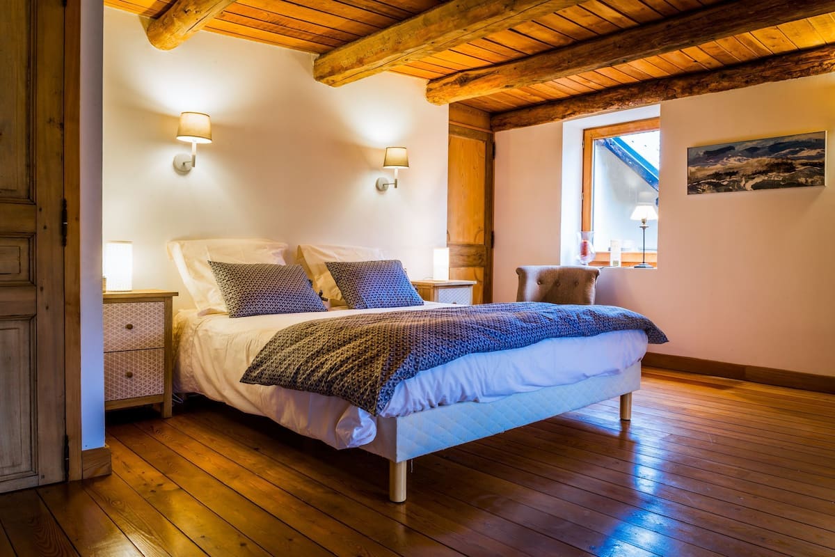 A bedroom with a wooden floor, a bed with patterned bedding, two wall-mounted lamps, and a window with a view.