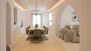 A modern dining area with a long table and chairs, a sofa, and a staircase. - Villa Opal (Mykonos)