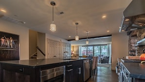 A modern kitchen with a large island, stainless steel appliances, pendant lighting, and a view to the living area. - Carriage House of New Hope (New Hope)