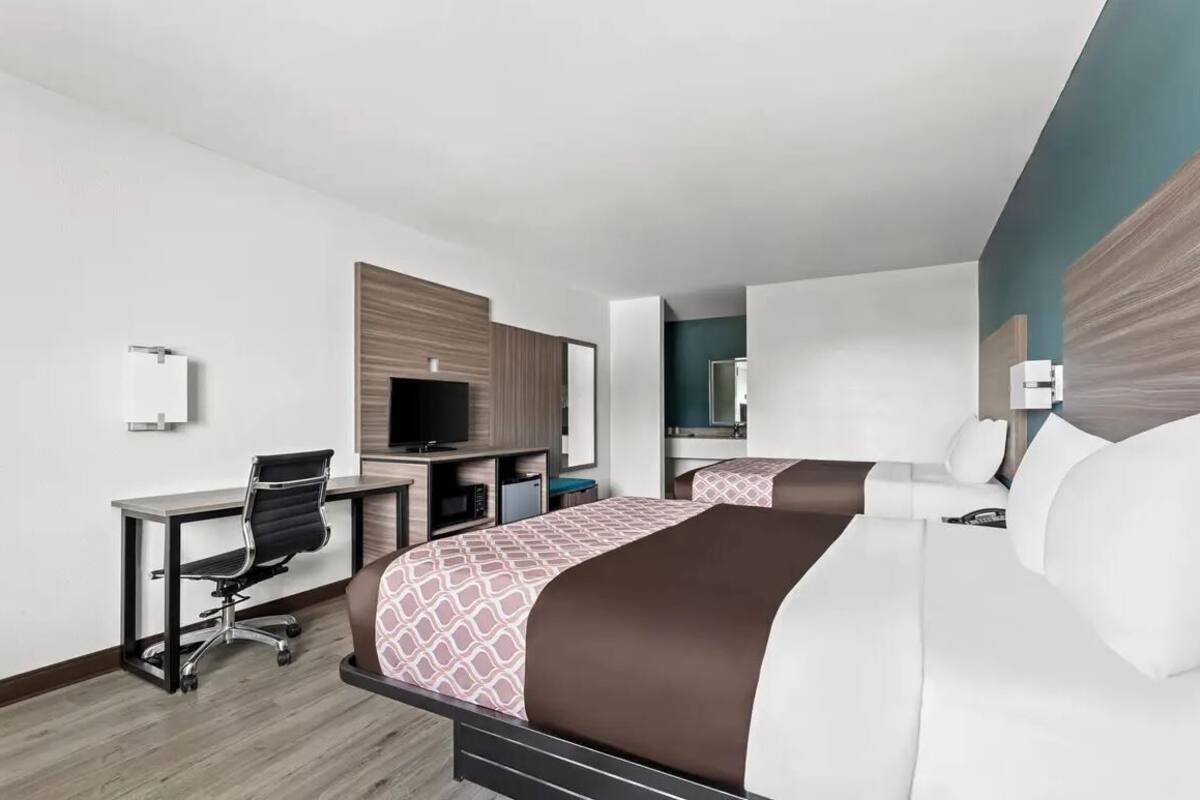 Quarto de hotel com duas camas, uma escrivaninha, uma cadeira, uma televisão e um espelho.