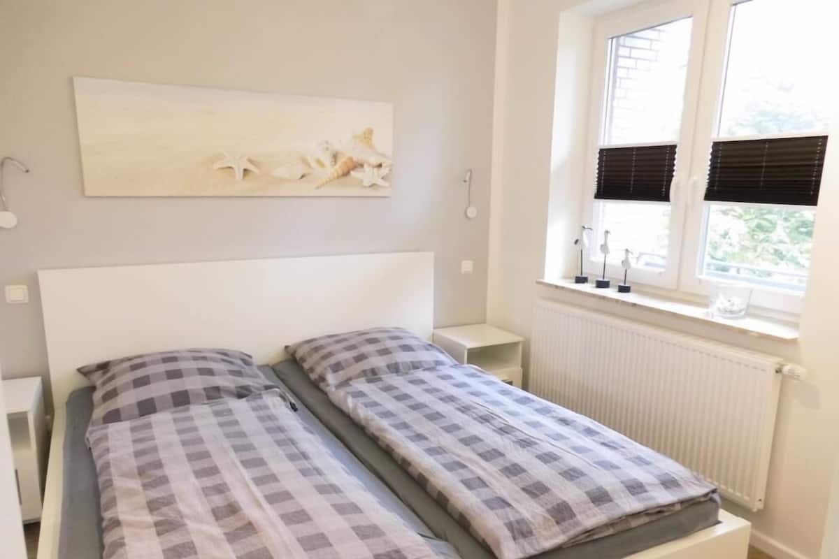 A double bed with a checkered bedspread, a bedside table, a wall-mounted lamp, and a window with blinds.