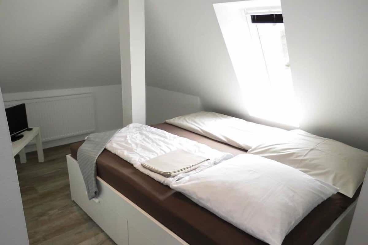 A small, neatly arranged bedroom with a single bed, a bedside table, and a window providing natural light.