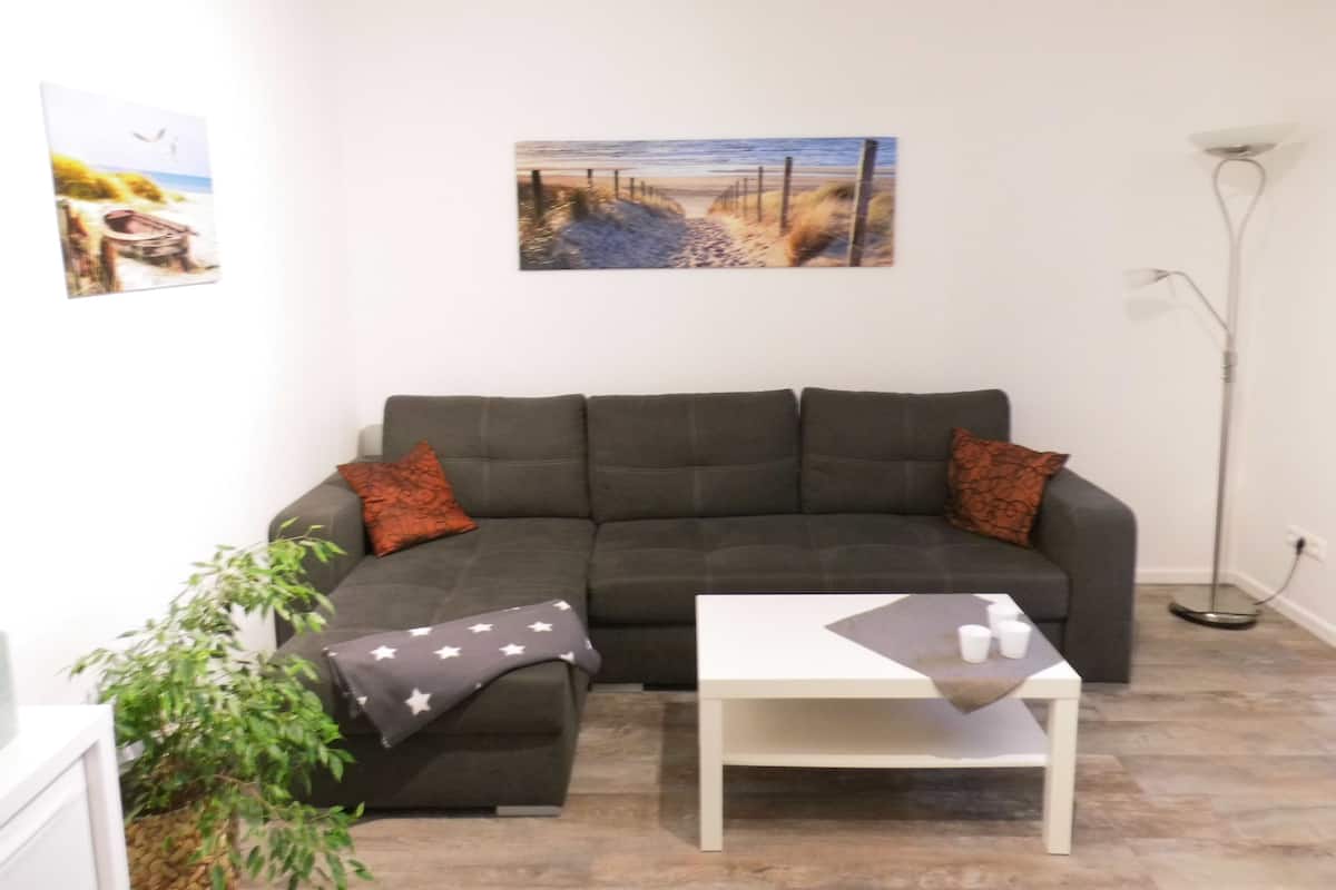 A living room with a dark green sofa, two brown pillows, a white coffee table, and a painting of a beach scene on the wall.