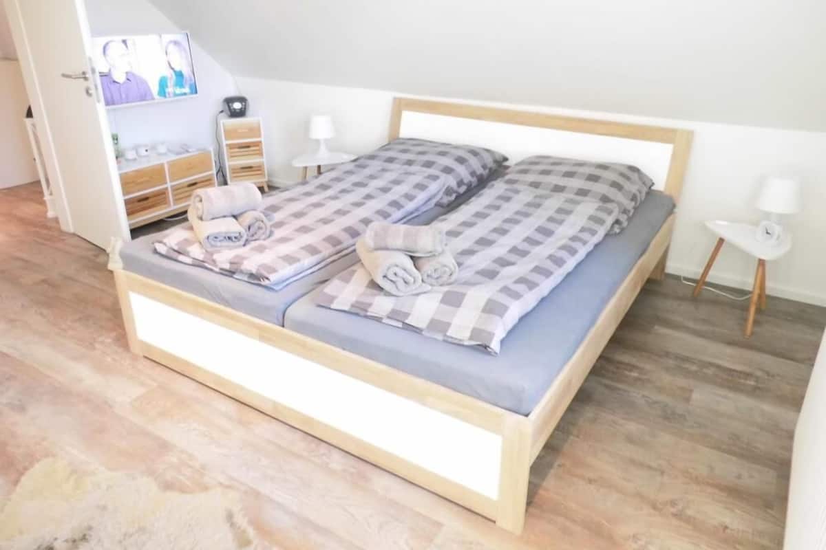 A bedroom with a wooden bed, checkered bedding, a bedside table with a lamp, and a TV on the wall.